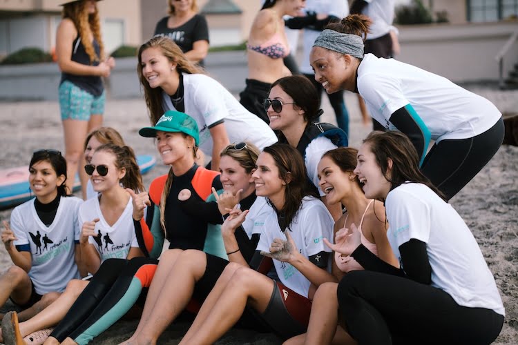 A group photo of Bethany Hamilton and many of the Beautifully Flawed attendees sitting on the beach smiling and giving shakas.