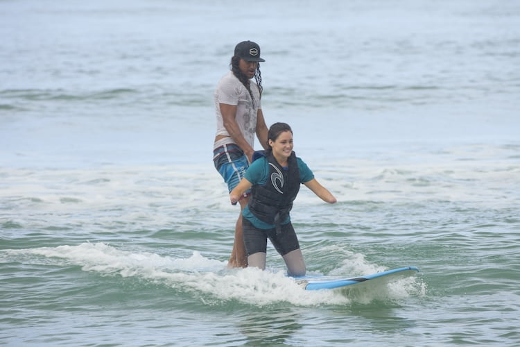 Quad amputee Amy stands on her surfboard and rides a waves with the help of her instructor.